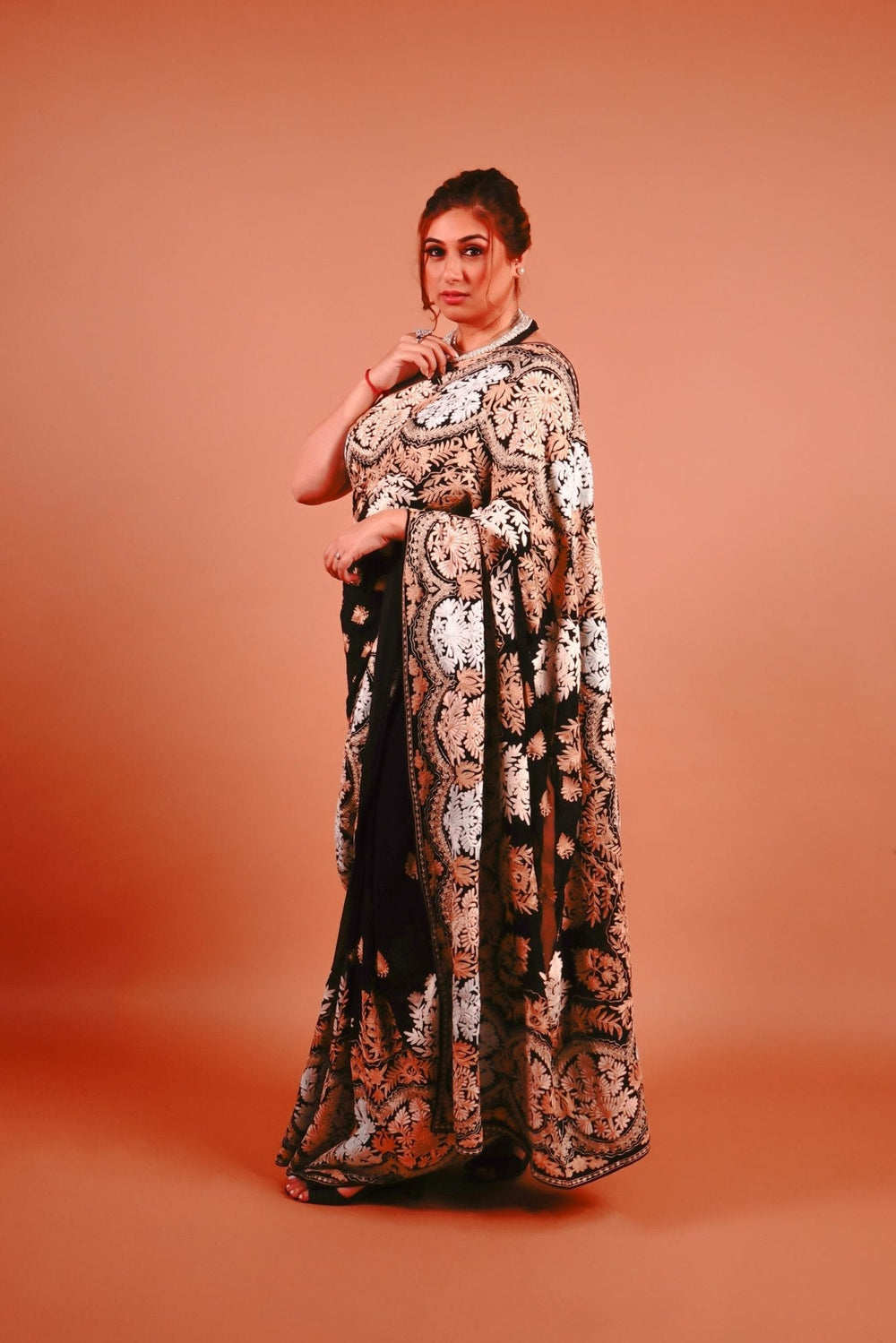 A woman standing in a pose wearing a black and gold Kashmiri embroidered saree with floral patterns.